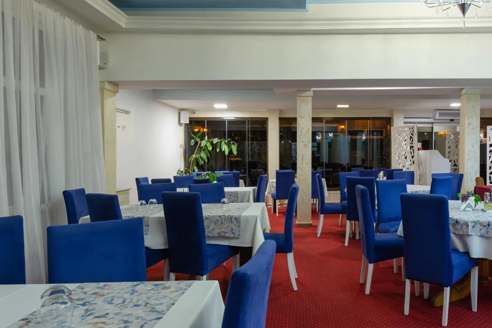 Salonul restaurantului cu scaune albastre tapițate, mese cu fețe de masă albe și mochetă roșie